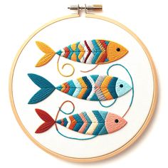 three colorful fish are on a white surface in an embroidery hoop, with thread spools