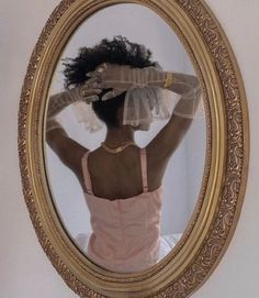 a woman in a pink dress looking at her reflection in a gold framed oval mirror