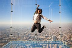 a woman is jumping in the air from a glass floor high above cityscape