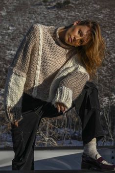 a woman sitting on the hood of a car wearing a sweater and black pants, with her hands in her pockets