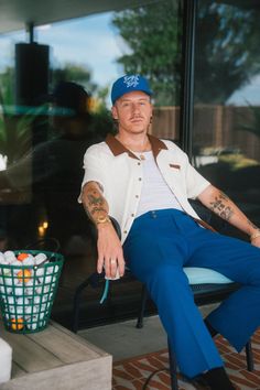 a man with tattoos sitting on a chair in front of a glass door wearing blue pants and a white shirt