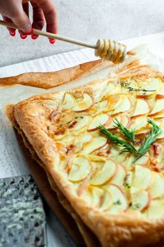 a person is holding a honey stick over a baked pastry with apples and rosemary on top