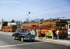 an old car is parked in front of the trolley cars on the street while people are waiting to board