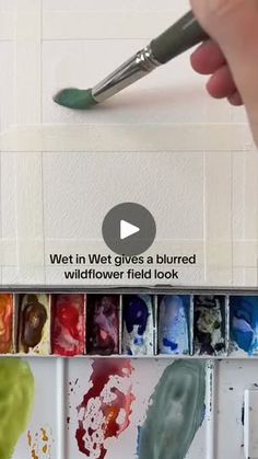 a person holding a paint brush in front of a wall filled with watercolors