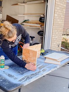 a woman is working on a piece of wood at a table in front of a garage