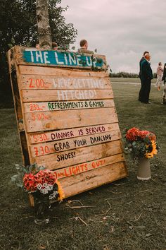 a wooden sign sitting on top of a grass covered field next to flowers and trees