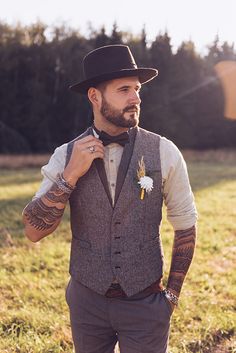 a man wearing a hat and vest with tattoos on his arm standing in the grass