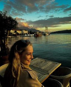 a woman sitting at a wooden table next to the water with boats in the background