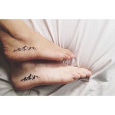 two feet with mountains tattoo on them sitting on a white bedding sheet in front of a pillow