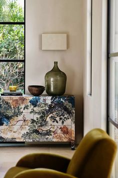 a green vase sitting on top of a wooden cabinet next to a yellow chair in front of a window