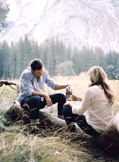 a man and woman sitting on a log in the grass with a mountain behind them