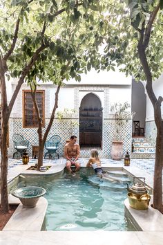 two people are sitting in the middle of a pool with trees and potted plants