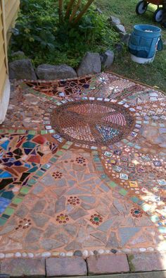 an outdoor walkway made out of various colored tiles and stones, with a wheelbarrow in the background