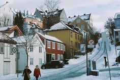 two people walking down a snowy street in the middle of some small town with tall buildings