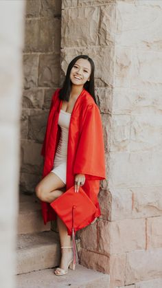 a woman in an orange graduation gown leaning against a stone wall and smiling at the camera