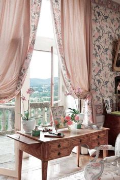 a desk in front of a window with pink curtains and flowers on the windowsill