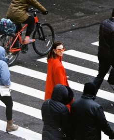 a woman in an orange coat is crossing the street with other people and bicycles behind her