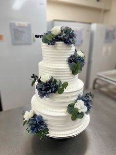 a white wedding cake with blue flowers and greenery on the top is sitting on a table