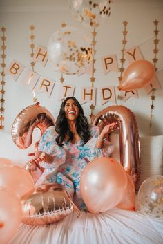 a woman sitting on top of a bed surrounded by pink balloons and gold foil letters