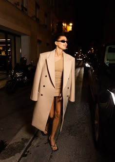 a woman is walking down the street at night with her coat over her shoulders and sunglasses on