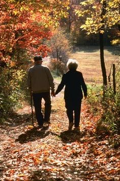 an elderly couple walking down a path in the woods with autumn leaves on the ground