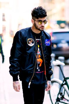 a man walking down the street wearing sunglasses and a jacket with patches on his sleeves