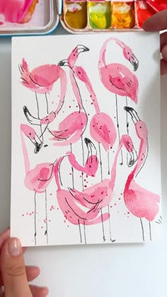 a person holding up a painting with pink flamingos on it in front of some watercolors