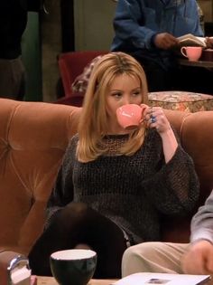 a woman sitting on a couch drinking from a pink coffee cup while another man sits in the background