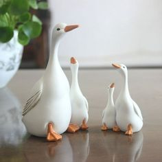 three white ducks standing next to each other on top of a wooden table in front of a potted plant