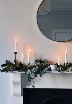 a mantle with candles and greenery in front of a round mirror on the wall