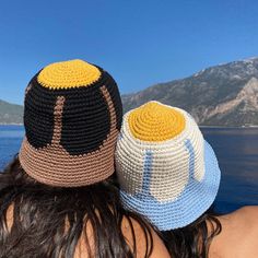 two people wearing crocheted hats looking out over the water at mountains and blue sky