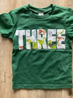 a green t - shirt with the word three printed on it, sitting on top of a wooden floor