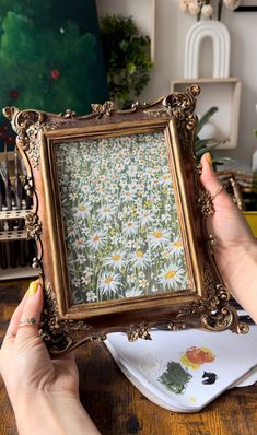 a person holding up an ornate frame with daisies on it in front of a painting