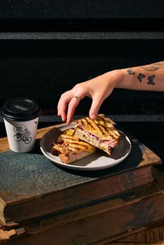 a person reaching for a sandwich on a plate with a cup of coffee next to it