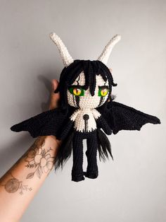 a hand holding up a knitted doll with green eyes and horns on it's head