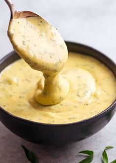 a wooden spoon scooping some yellow sauce out of a black bowl with green leaves on the side