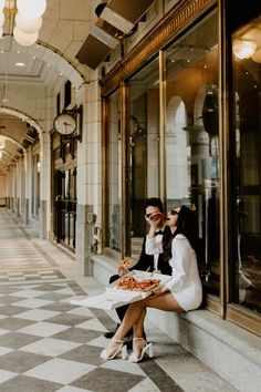 two women sitting on the side of a building, one holding a plate of pizza