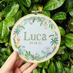 someone is holding up a cross - stitch hoop with the name lucia on it in front of some green leaves