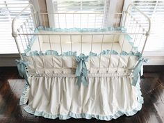 a white crib with blue ruffles and pillows on the bottom, in front of a window