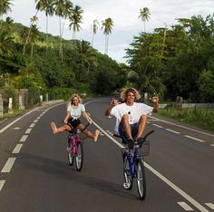 two people riding bikes on the road with palm trees in the background