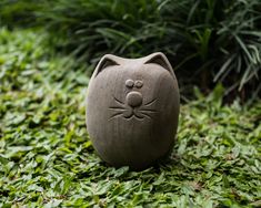 a clay cat head sitting on top of green grass next to bushes and plants in the background