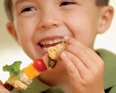 a young boy eating a piece of fruit and veggie skewer while smiling at the camera