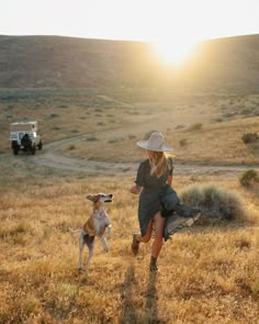 a woman in a hat and dress running with a dog on a grassy field at sunset