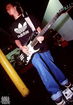 a young man playing an electric guitar in front of a yellow pole and some people