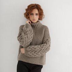 a woman in a sweater leaning against a wall with her hand on her chin and looking at the camera