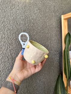 a hand holding a ceramic cup with a tennis racket in it and a mirror behind it