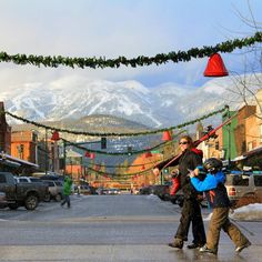 two people walking down the street with snow covered mountains in the background and lights strung over them