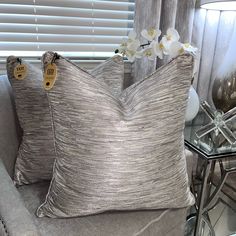 two decorative pillows on a couch in front of a window with white flowers and glass side table