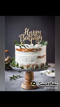 a white cake with greenery on top is sitting on a wooden stand and has a happy birthday sign