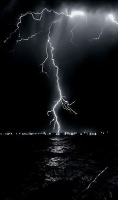 a black and white photo of lightning over the ocean at night with bright lights in the sky